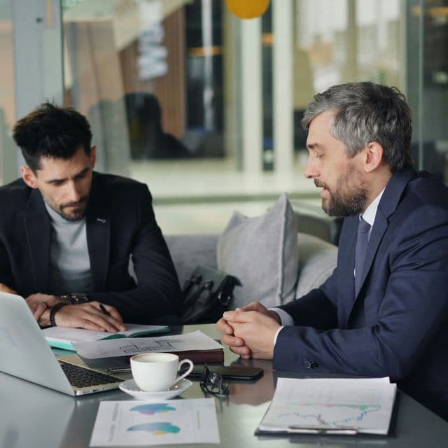 two people all on laptops, two men listen to person talking in a board meeting